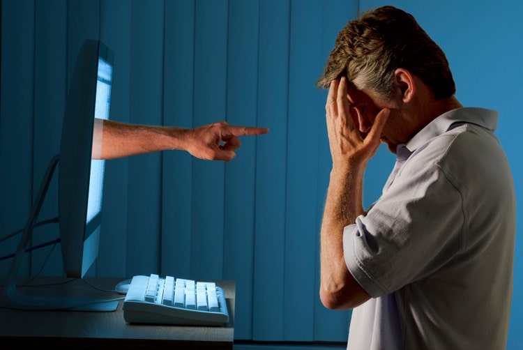 Severely distraught young man sitting in front of a computer with a judgmental hand pointing at him from within the computer monitor