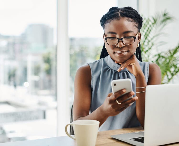 Young black businesswoman using a cellphone while working on a laptop in an office alone