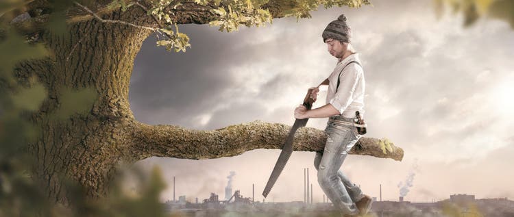 A man is sitting on a branch he is about to cut off. The background shows an industrial area which is polluting the surroundings. Thus the image conveys a secondary meaning towards environmental pollution.