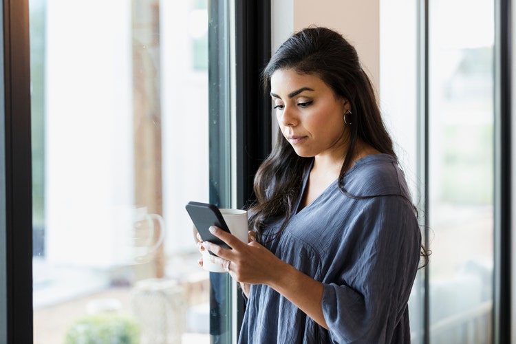 A thoughtful woman reads a text message while standing next to a window in her home.