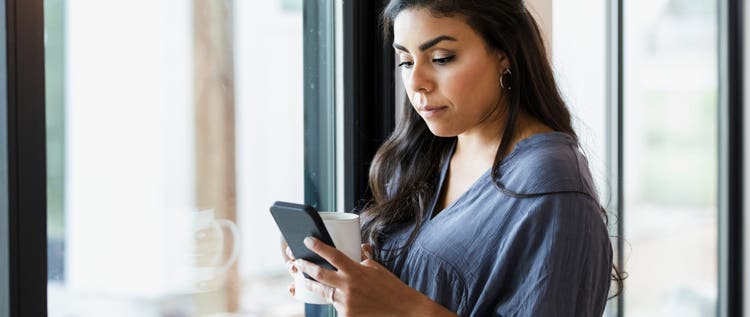 A thoughtful woman reads a text message while standing next to a window in her home.