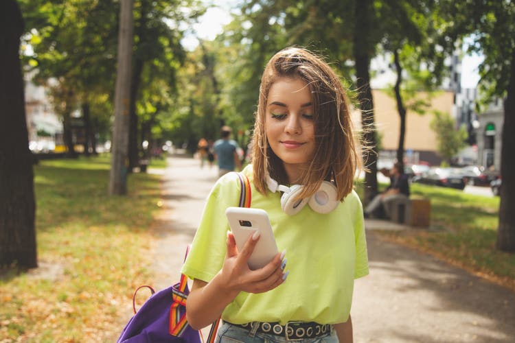 Happy beautiful young high school girl with white smart phone outdoors on sunny summer day texting and smiling.