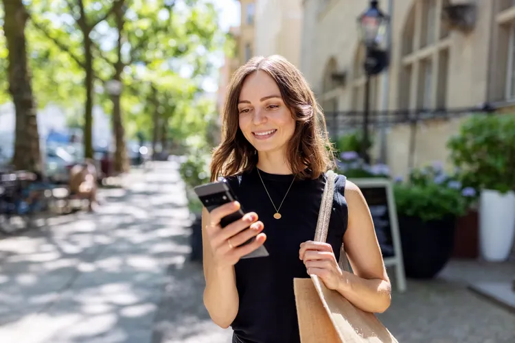 Smiling woman checking her phone while walking along a tree-lined city street