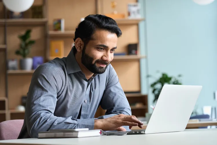 Man smiling while working on a laptop in a bright home office